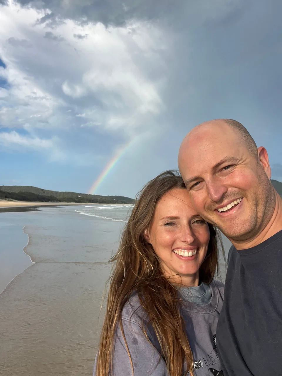 Kyle and Kelly smiling together on the beach beneath a rainbow