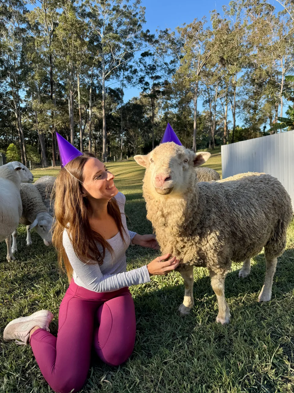 Kelly crouching beside a sheep, both wearing party hats